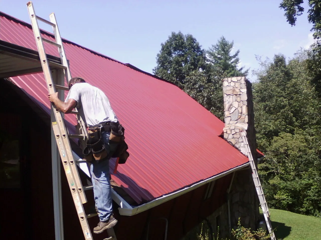 Roofer on a ladder beside a red metal roof and stone chimney — Soffit & Fascia Repair by Roofing Brothers in Canisteo